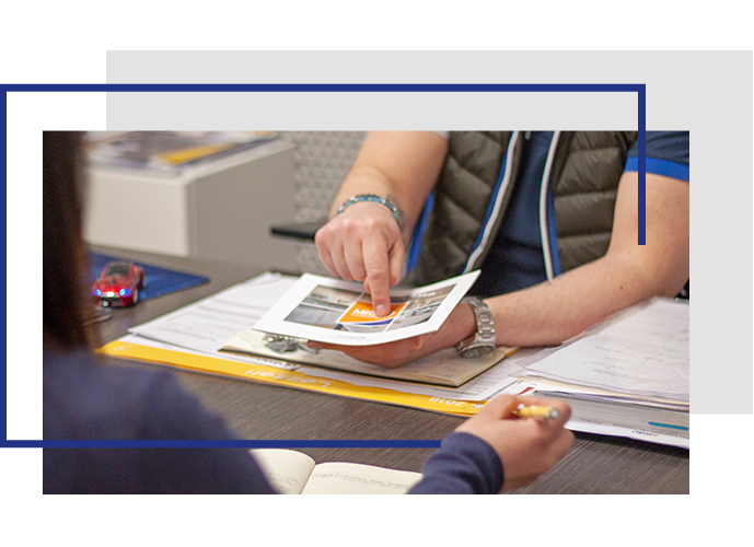 Su una scrivania, due braccia maschili mostrano una brochure ad una donna seduta di spalle, lei tiene una penna gialla nella mano destra, lui con la destra indica la brochure che sostiene con la sinistra