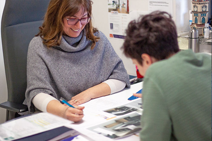Due donne sorridono guardando un progetto sulla scrivania, una porta gli occhiali e ha in mano una penna, l’altra è di spalle e osserva i fogli